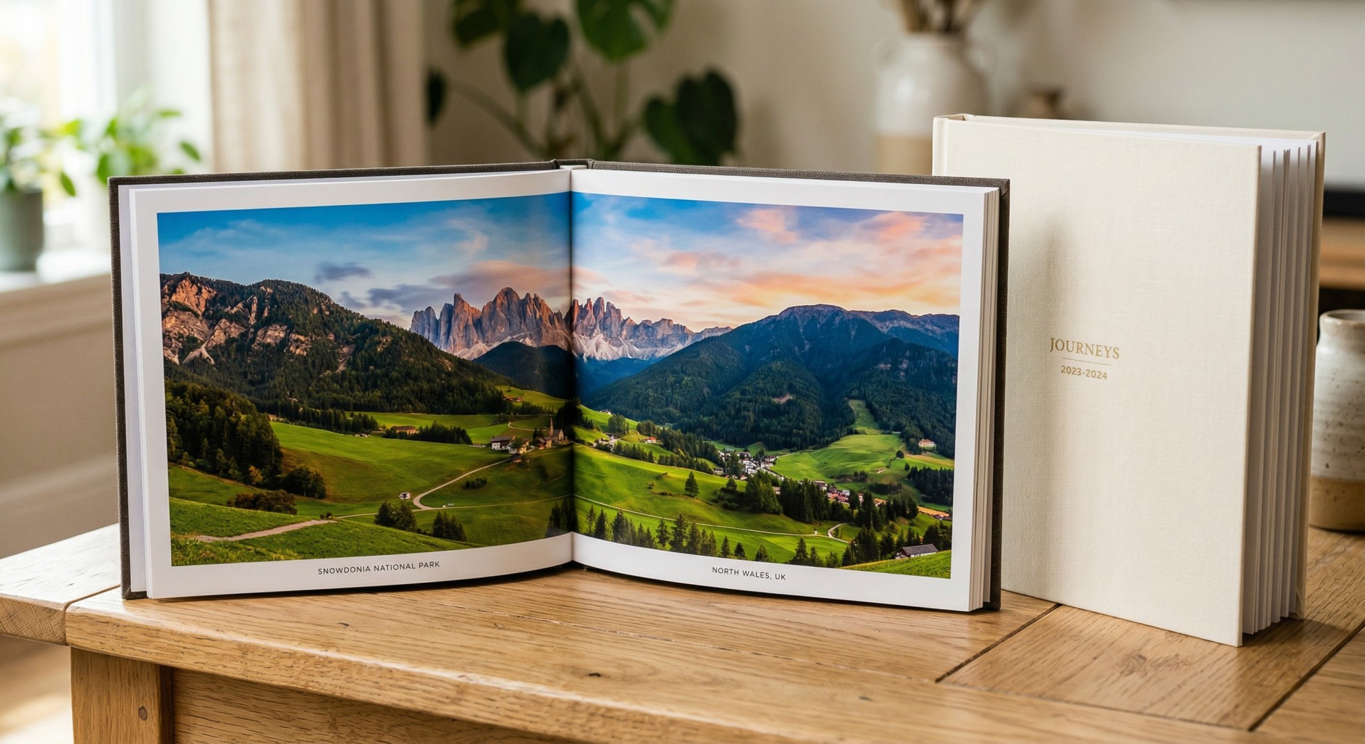 Open hardcover photo book on a wooden table showing a landscape photograph spread on glossy pages, with a closed cream-covered photo book beside it