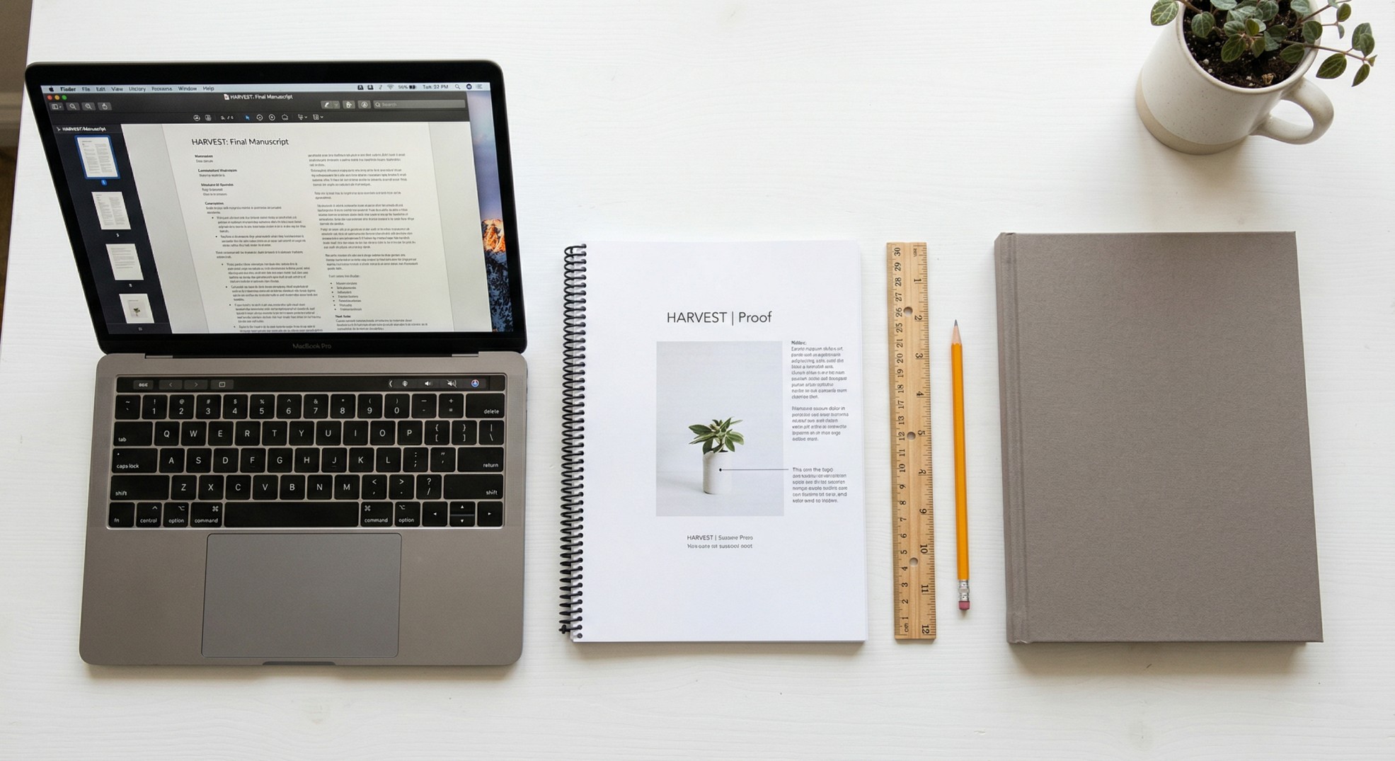 Flat-lay overhead photo showing the hardcover creation workflow - laptop with PDF manuscript, spiral-bound proof copy, and finished gray hardcover book with a ruler and pencil