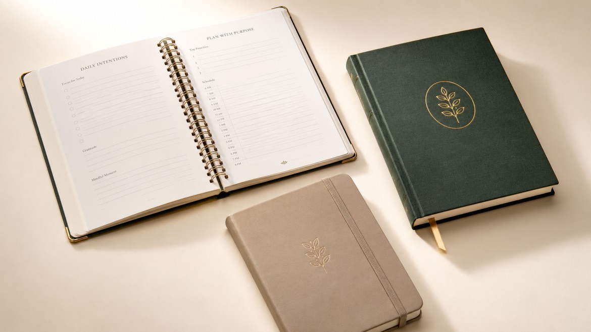 Three premium custom journals on a cream background: an open spiral-bound planner, a foil-stamped hardcover journal, and a journal with an elastic closure and ribbon bookmark